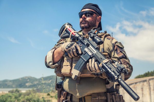 Private military company mercenary, brutal looking special forces fighter in battle uniform and plate carrier, wearing radio headset and sunglasses, holding service rifle in hands, ready to fight